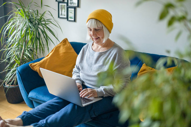 Χαλαρωμένη μεσήλικη 50άρα γυναίκα σε casual στυλ φοράει κάθεται στο σπίτι στο σαλόνι, χρησιμοποιώντας το laptop, αγοράζοντας online, εργασία από το σπίτι  - Φωτογραφία, εικόνα