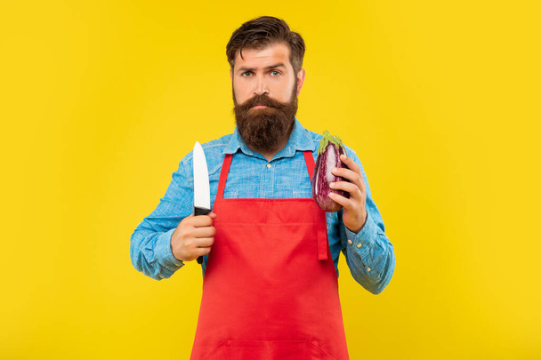Serious guy in apron holding cooks knife and eggplant yellow background, cook - Fotó, kép