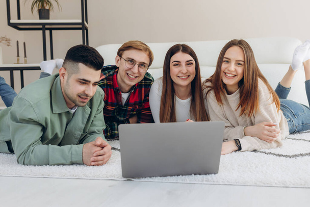 Happy teenage friends having fun spending time together watching laptop lying on floor near each other in design comfortable living room - Foto, imagen