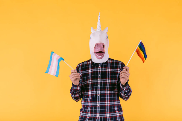 Personne vêtue d'un masque de licorne avec une chemise à carreaux tenant un drapeau de la communauté transsexuelle et lgtbq, sur un fond jaune. Concept de fierté gay, transsexualité, genre, binaire et lesbienne - Photo, image