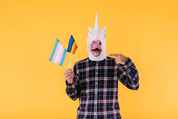 Person dressed in a unicorn mask with a plaid shirt holding a flag of the transsexual and lgtbq community, on a yellow background. Concept of gay pride, transsexuality, gender, binary and lesbian - Foto, Imagem