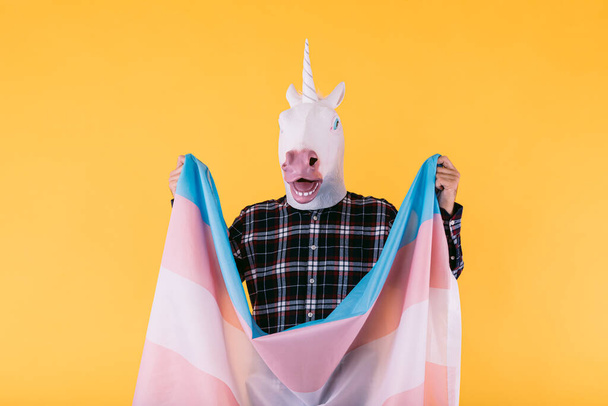 Person dressed in a unicorn mask with a checkered shirt holding the flag of the transsexual collective, on a yellow background. Concept of gay pride, transsexuality, and lgtbq rights. - Foto, Imagen