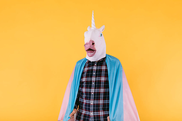 Persona vestida con una máscara de unicornio con una camisa a cuadros que sostiene la bandera del colectivo transexual, sobre un fondo amarillo. Concepto de orgullo gay, transexualidad y derechos lgtbq. - Foto, imagen