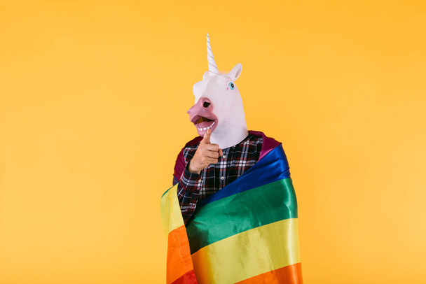 Persona vestida con máscara de unicornio con camisa a cuadros que sostiene la bandera del arco iris lgtbq, sobre fondo amarillo. Concepto de orgullo gay, transexualidad y derechos lgtbq. - Foto, imagen