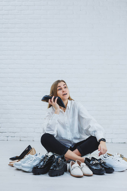 Happy beautiful young woman with a smile in a fashionable outfit with a white shirt and black trousers sits on the floor among the shoes and chooses a new pair of shoes for herself - Foto, imagen