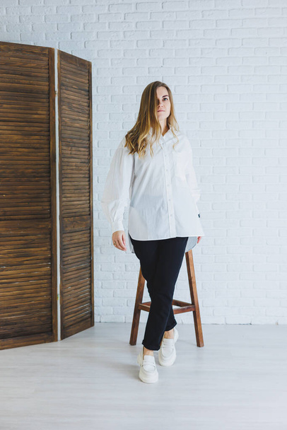 A young woman in black pants and a white shirt against a white brick wall, a stylish modern girl in white leather loafers - Foto, Bild