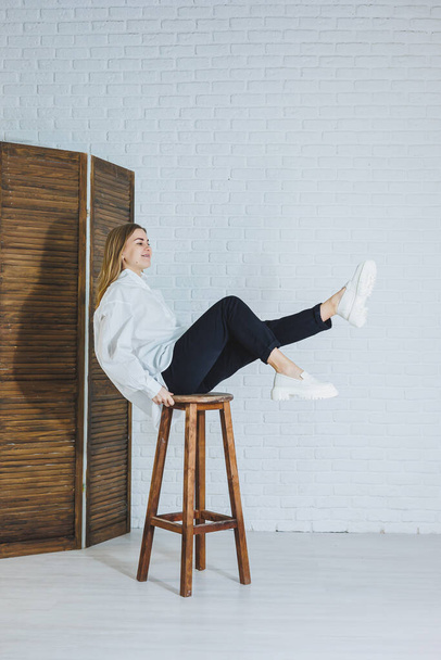 A beautiful woman is wearing a white cotton shirt and black pants. Cheerful emotional girl sits on a wooden chair against a white brick wall. - Photo, Image