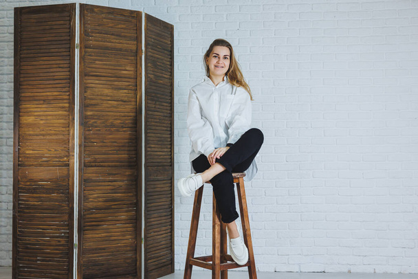 A beautiful woman is wearing a white cotton shirt and black pants. Cheerful emotional girl sits on a wooden chair against a white brick wall. - Фото, изображение