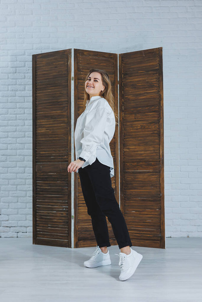 A beautiful stylish woman is wearing a white cotton shirt and black pants. Cheerful emotional girl in white sneakers - Fotografie, Obrázek