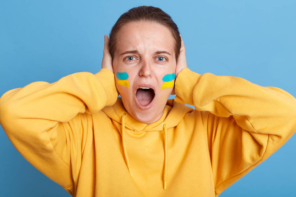 Desperate woman in yellow hoodie with Ukrainian flag on cheeks, posing isolated over blue background, standing and covering her ears, don't want listen about war in Ukraine, screaming. - Фото, зображення