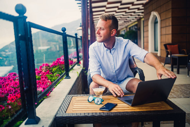 Успешный бизнесмен сидит за столом с ноутбуком, работает в отпуске. Отпуск и удаленная работа. Парень сидит в ресторане на террасе с панорамным видом - Фото, изображение