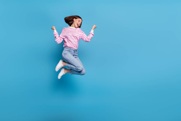 Photo of excited lucky woman wear striped sweater rising fists jumping high empty space isolated blue color background - Фото, изображение