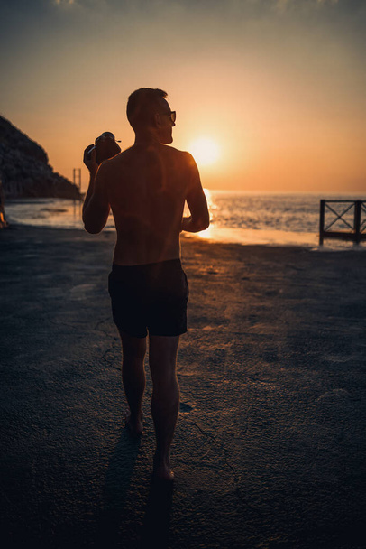 Jeune photographe masculin fait des photos de la mer au coucher du soleil tout en se tenant sur le rivage. Coucher de soleil. Concentration sélective. Photographies touristiques du paysage marin - Photo, image