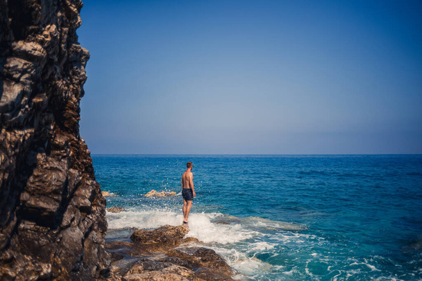 Een jongeman staat op de rotsen met uitzicht op de open Middellandse Zee. Een kerel op een warme zomer zonnige dag kijkt naar de zeewind - Foto, afbeelding
