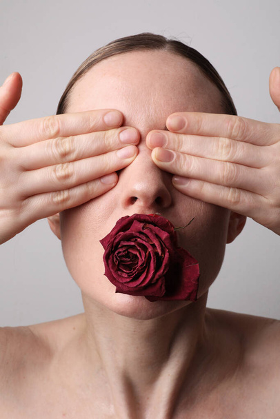 Vertical portrait of young woman with dry rose covering her eyes on white wall. - Фото, изображение