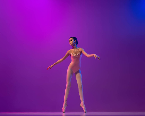 Studio shot of beautiful school age girl, ballet dancer dancing isolated on purple background in neon light. Art, grace, beauty, ballet school concept - Foto, Imagem
