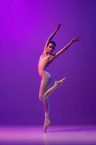 Studio shot of beautiful school age girl, ballet dancer dancing isolated on purple background in neon light. Art, grace, beauty, ballet school concept - 写真・画像