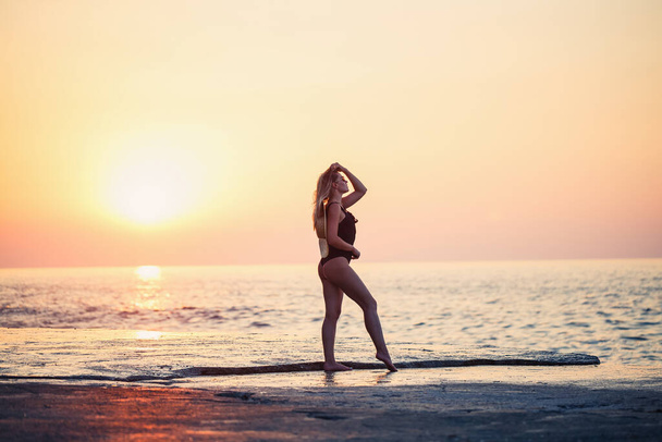 Kaunis nainen mustassa uimapuvussa meren rannalla, hymyilevät kasvot olkihattu olkapäillään. Kultainen auringonlasku valo - Valokuva, kuva