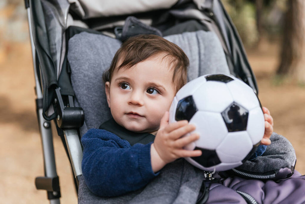 Маленька дитина тримає футбольний м'яч, сидячи в колясці на відкритому повітрі в парку
. - Фото, зображення
