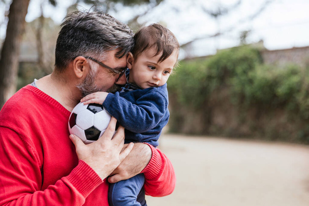 Homem segura seu pequeno filho e uma bola de futebol enquanto jogam juntos ao ar livre no parque. - Foto, Imagem