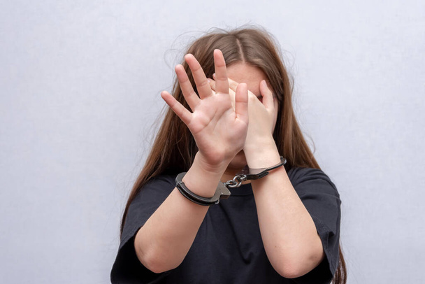 Een meisje met handboeien verbergt haar gezicht op een grijze achtergrond, close-up. Jeugdige delinquent in zwart T-shirt, strafrechtelijke aansprakelijkheid van minderjarigen. - Foto, afbeelding