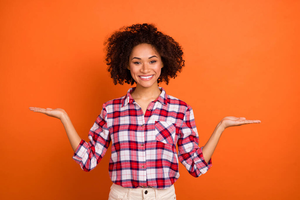 Photo of young curly lady hold empty space wear plaid shirt isolated on orange color background - Foto, Bild