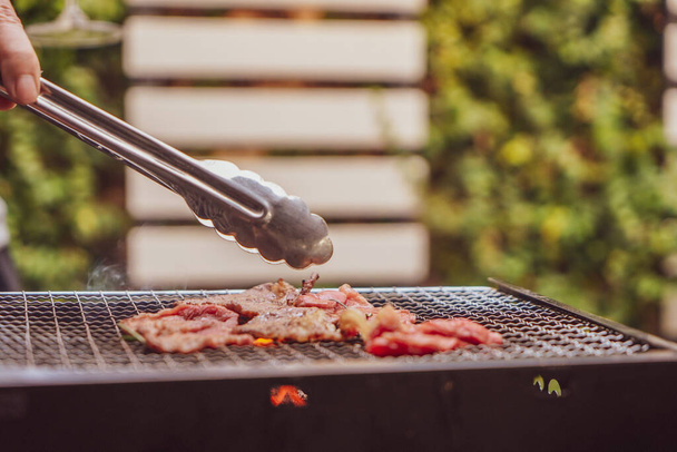 Close up view of pieces of sliced beef on charcoal grill at Outdoor party. Human's hands using meat tongs to grill Multiple slices of premium meat on the stovetop. Home made barbecue beef grill smoke. - Foto, imagen