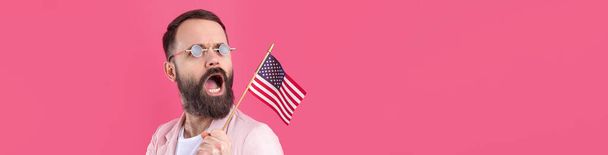 Retrato de um jovem satisfeito com uma barba com uma bandeira americana em um fundo de estúdio vermelho. Grande patriota dos EUA e defensor da liberdade. - Foto, Imagem