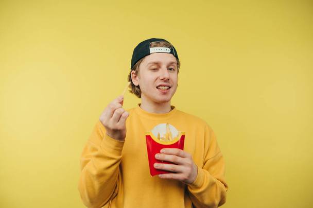 Positieve jongeman met frietjes in zijn handen staat op een gele achtergrond en kijkt met een glimlach op zijn gezicht naar de camera. - Foto, afbeelding