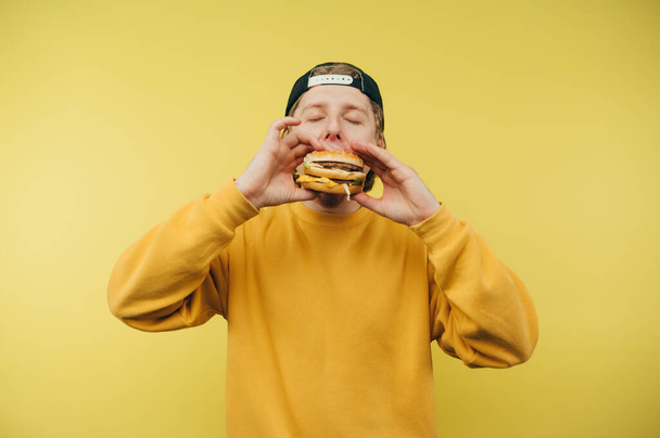 Hungry young man in a cap and casual clothes eats an appetizing burger on a yellow background, bites with his eyes closed. - Foto, Imagen