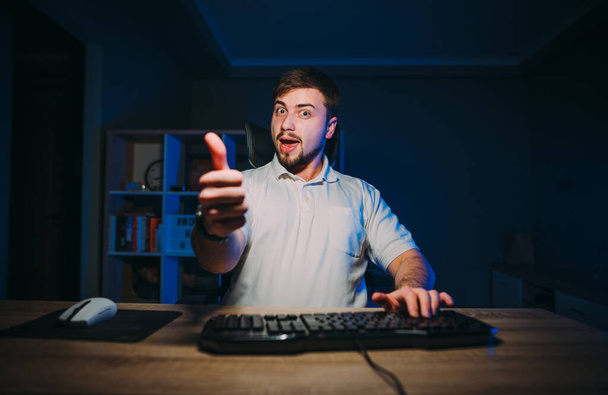 Giovane lavoratore maschio positivo seduto di notte al lavoro al computer, guardando la fotocamera e mostrando un gesto che gli piace. - Foto, immagini