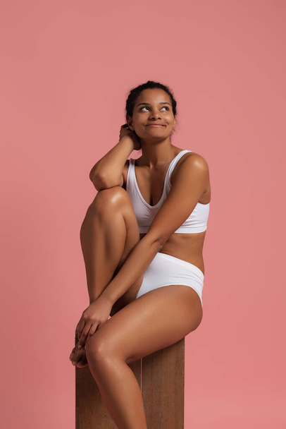 Portrait of young beautiful slim woman sitting, posing in white cotton underwear isolated over pink studio background. Bosy positivity concept - Φωτογραφία, εικόνα