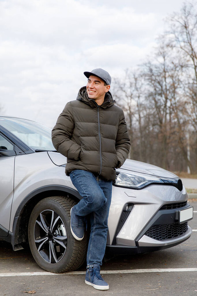 A young cheerful guy in a jacket and a cap stands near a new car - Foto, imagen