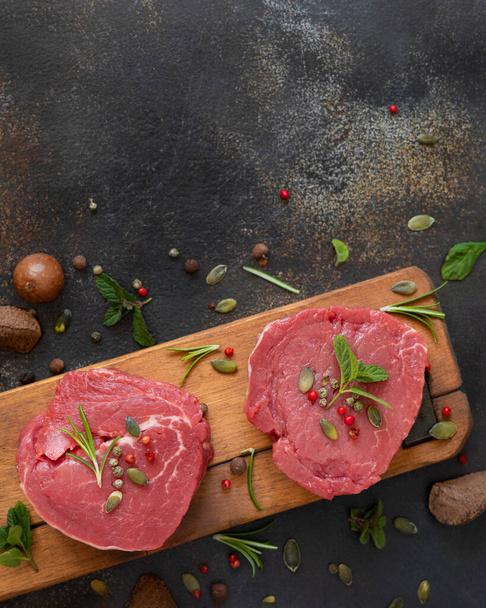 Raw beef fillet steaks on wooden board on dark table close up with copy space. Portioned filet mignons with spices and herbs. Concept of summer food - 写真・画像