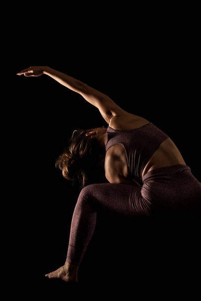 Fit woman practicing yoga poses. Side lit half silhouette girl doing exercise in studio against black background. - Фото, зображення