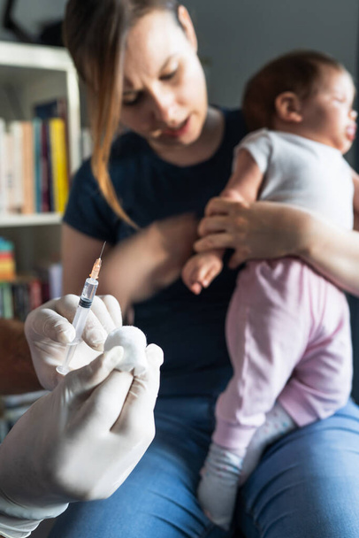 Close up on hand of unknown man doctor wearing protective medical gloves and holding a syringe preparing injection shot with vaccine for small caucasian baby mother hold her child for vaccination - Photo, Image