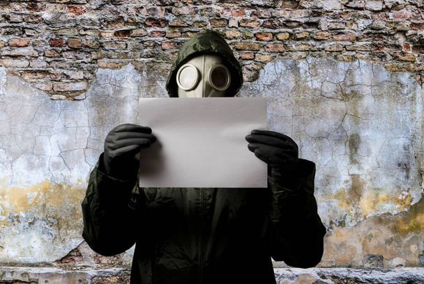 Man wearing a gas mask on his face and holding white paper, the wall behind him with copy space. Danger of chemical weapon. - Foto, afbeelding