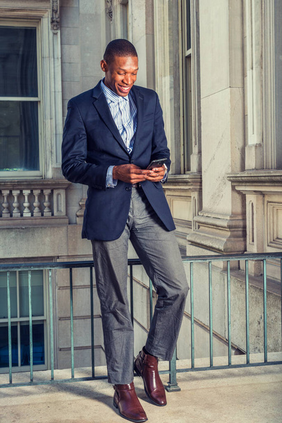 African American businessman in New York. Wearing black blazer, gray pants, brown leather shoes, a young guy standing in vintage style office, looking down, smiling, reading messages on mobile phone - Photo, Image