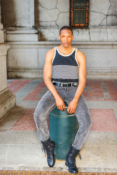Man casual street fashion. Wearing black, white striped tank top, jeans, leather boots, a young African American guy sitting on street in New York, relaxing, thinking, lost in thought - Foto, Imagem