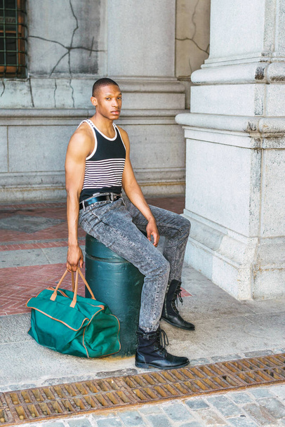 Moda callejera casual de hombre. Vestido negro, camiseta blanca a rayas, jeans, botas de cuero, bolsa de mano verde, un joven afroamericano sentado en la calle en Nueva York, esperando, relajándose - Foto, imagen