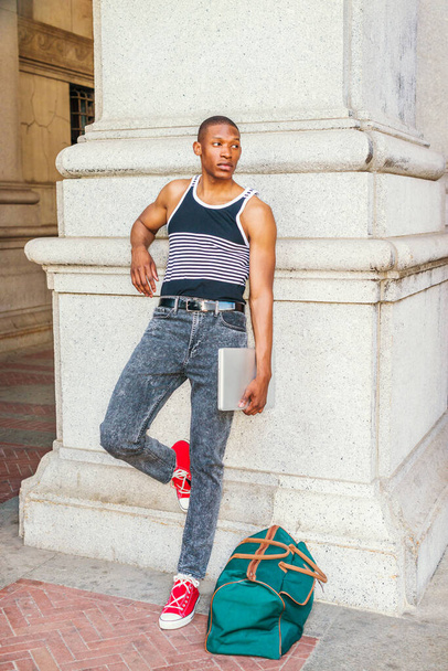 College student traveling in summer in New York. Wearing black, white striped tank top, jeans, red sneakers, carrying green bag, holding laptop computer, a black guy standing against column on street. - Photo, Image