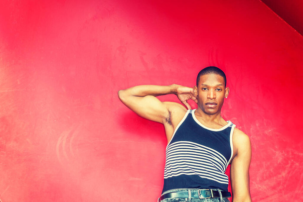 Summer Heat. Man casual fashion. Wearing black, white striped tank top,  raising arm, hand touching shoulder, young African American guy leaning against red metal wall, seriously looking at you - Fotó, kép