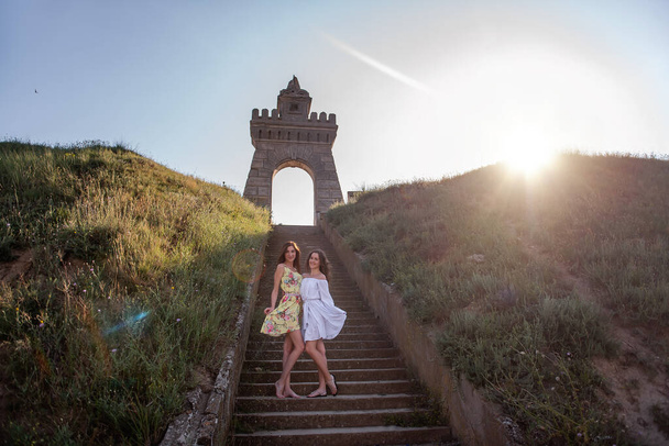 Two young women in summer dresses hold hands, hug each other against the background of a staircase with a vintage arch. Beautiful girls sisters travel together on a weekend at sunset. - Valokuva, kuva