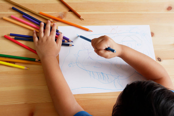 Słodki azjatycki chłopiec rysunek szczęśliwej rodziny i domu. z kolorowymi ołówkami i białym papierem na drewnianym stole, widok z góry - Zdjęcie, obraz