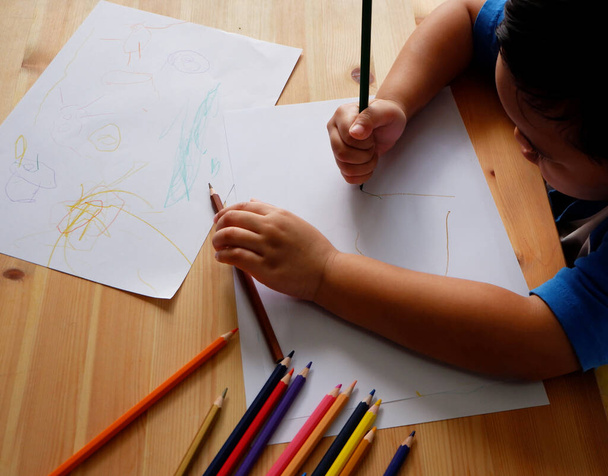 Cute Asian boy drawing with color pencils and white paper on wooden table - 写真・画像