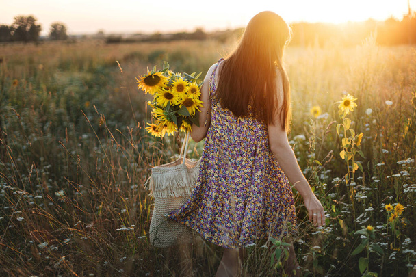 Mulher bonita reunindo girassóis em luz quente do pôr do sol no prado de verão. Momento atmosférico tranquilo no campo. Elegante jovem fêmea em vestido floral segurando girassóis no campo da noite - Foto, Imagem