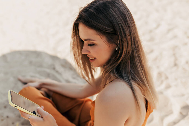 Top view kuva rakastettava viehättävä brunette tyttö lepää ulkona älypuhelimella. Euroopan tyttö istuu hiekalla aurinkoinen lämmin päivä ja vieritys älypuhelin auringonpaisteessa. Laadukas kuva - Valokuva, kuva