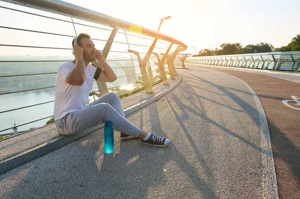 Ελκυστικός χαλαρός νεαρός μυώδης αθλητής που βάζει ακουστικά και ακούει μουσική, κάθεται σε μια γέφυρα και απολαμβάνει τα υπόλοιπα μετά την πρωινή προπόνηση. Όμορφες ακτίνες του ήλιου πέφτουν στο διάδρομο - Φωτογραφία, εικόνα