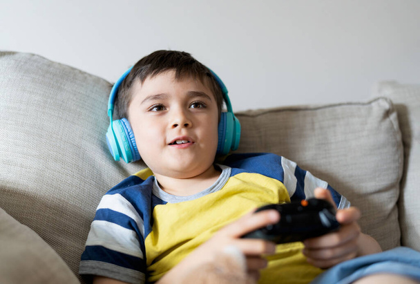 Kulaklık takan ve video oyunu oynayan portre çocuğu. Çocuk evde oyun konsolu tutuyor, genç çocuk kanepede oturuyor ve hafta sonları tek başına eğleniyor. - Fotoğraf, Görsel