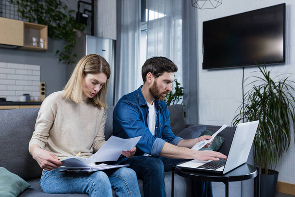 Young family, husband and wife, reviewing their bills, loan and mortgage agreements, sitting on the couch at home, working with documents on a laptop, embarrassed, upset - Photo, Image